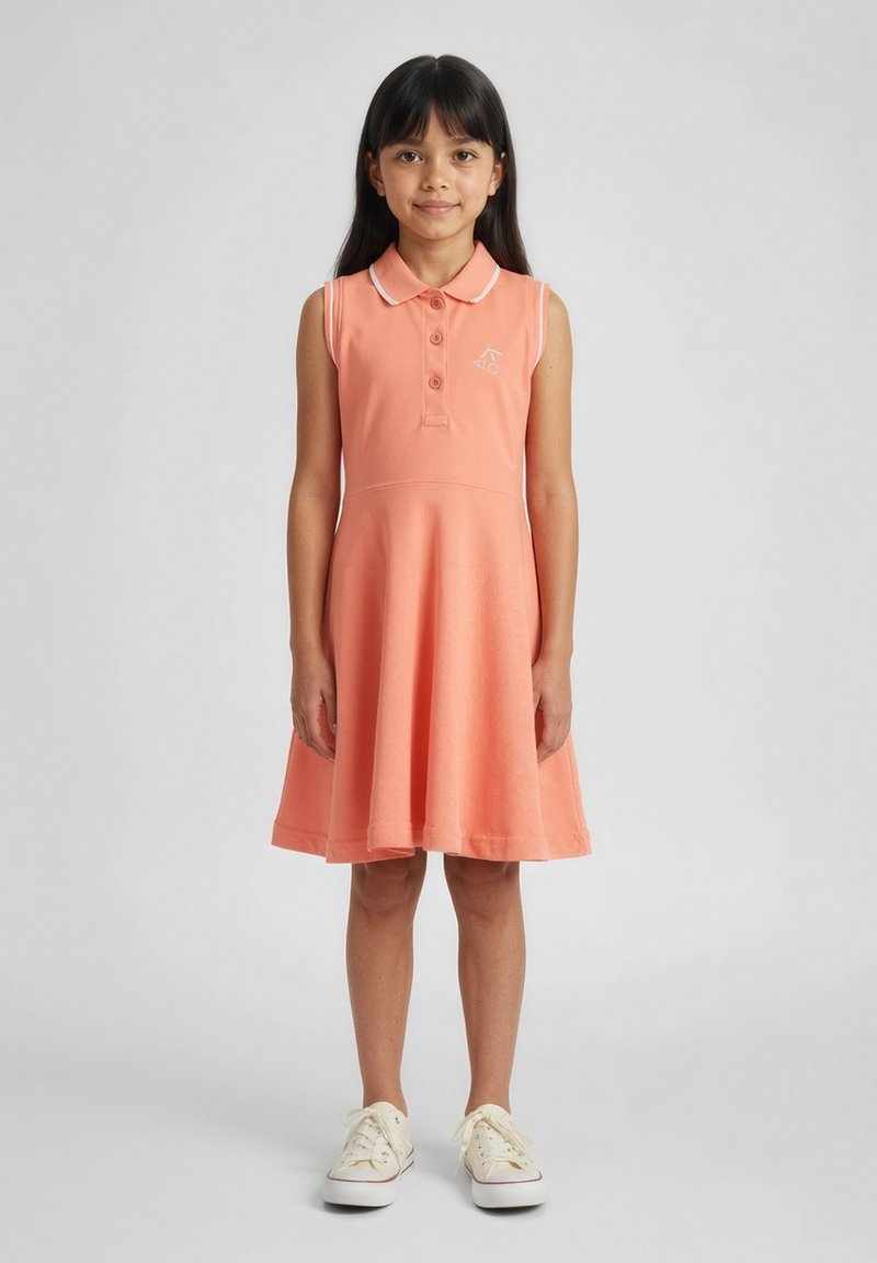 Młoda dziewczyna stoi twarzą do przodu, ubrana w bezrękawnikową brzoskwiniową sukienkę z kołnierzykiem i białe trampki na jednolitym tle.