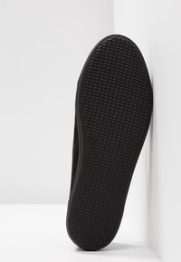 Semelle d'une chaussure noire avec motif de bande de roulement en zigzag, taille 37, positionnée verticalement contre des murs blancs en angle.