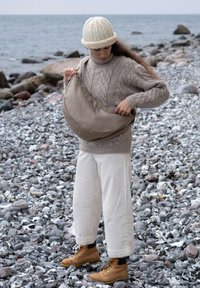 Borsa a tracolla beige di forma rettangolare, realizzata in materiale liscio, indossata da una persona con un maglione beige a trecce e pantaloni bianchi larghi, su una spiaggia rocciosa.
