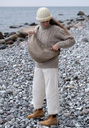 Bolso beige de hombro con forma rectangular, hecho de un material suave, llevado por una persona con un suéter de punto beige y pantalones blancos anchos, en una playa rocosa.
