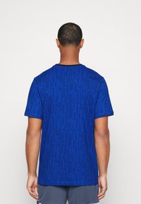 Modrá texturovaná tričko s žebrovaným vzorem a krátkým rukávem. Má kulatý výstřih a volný střih. Zadní pohled, minimalistický design.