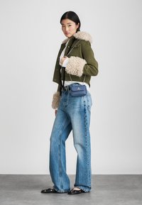 Γυναίκα που στέκεται φορώντας τζάκετ με πράσινη γούνα, λευκό μπλουζάκι, φαρδιά μπλε τζιν, μαύρες φλατ και μικρή μπλε τσάντα χιαστί, σε γκρι δάπεδο και λευκό φόντο.