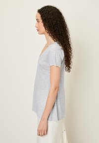 Šviesiai pilkas trumpas rankovėmis marškinėliai su V formos iškirpte, minkšta tekstūra, laisvas fitas ir apvaliu apačiu, dėvimi su šviesios spalvos sijonu.