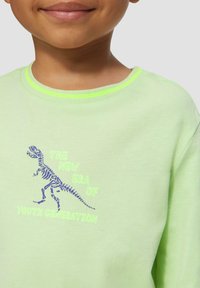 Hellgrüner Sweatshirt mit Rundhalsausschnitt, versehen mit einem blauen Dinosaurierskelett-Graphic und dem Text "DIE NEUE ÄRA DER JUGENDGENERATION."