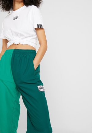 Kvinde iført en hvid Adidas cropped T-shirt med sorte striber og to-farvede grønne Adidas træningsbukser, hænderne i lommerne, mod en ensfarvet baggrund.