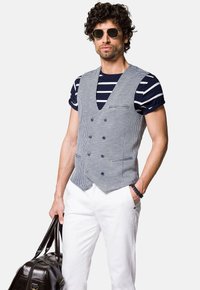 Szary kamizelka w kratę houndstooth z granatowymi guzikami na granatowej i białej pasiastym koszuli; białe spodnie i brązowa skórzana torba. Noszone okulary przeciwsłoneczne.