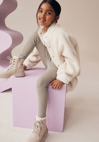 Veste en fausse fourrure blanche, leggings côtelés gris clair, bottes beige à lacets avec bord en fourrure, assise sur un cube rose, fond neutre.