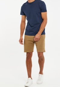Homme portant un t-shirt bleu marine, un short texturé couleur beige et des baskets blanches, debout devant un fond blanc uni.