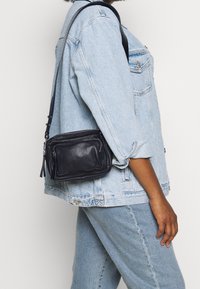 Lille sort læder crossbody taske med lynlåslommer, tekstureret finish og en aftagelig rem, vist mod en lys baggrund.