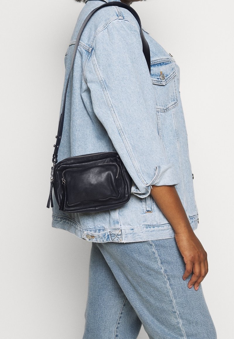 Lille sort læder crossbody taske med lynlåslommer, tekstureret finish og en aftagelig rem, vist mod en lys baggrund.