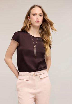 Junge Frau mit langen, welligen blonden Haaren, die eine dunkelbraune kurzärmelige Bluse, hellrosa Hose mit hoher Taille, eine goldene Halskette und Ohrringe trägt.