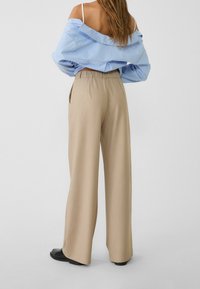 Pantalon beige à jambes larges avec une finition texturée, doté d'une taille haute et de poches latérales. Associé à une chemise oversize rayée bleue.