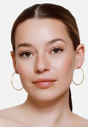 Młoda kobieta o brązowych oczach i włosach zaczesanych do tyłu, nosząca duże złote kolczyki koła, spokojnie patrzy przed siebie na jednolitym tle.