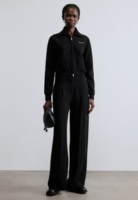 Fekete cipzáras pulóver két első zsebbel, széles szárú fekete nadrággal párosítva. A modell egy kis fekete kézitáskát tart, semleges háttér előtt áll.