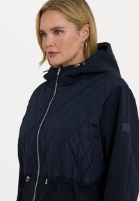Marineblå hettegenser har en quiltet overdel, elastisk liv og glidelåslommer. Materialet ser glatt ut med en myk tekstur og en subtil glans.