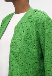 Vihreä pitsi-bomber-takki, jossa on monimutkaisia leikattuja kuvioita, ribbireunaiset kaulus ja hihansuut sekä vetoketjukiinnitys valkoisen paidan päällä.