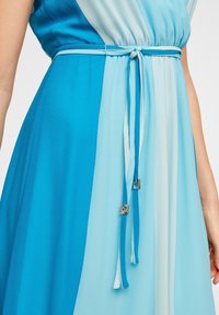 Robe bleu et bleu clair avec une taille cintrée, dotée d'un lien en tissu et d'accents métalliques. Le matériau satiné tombe doucement.