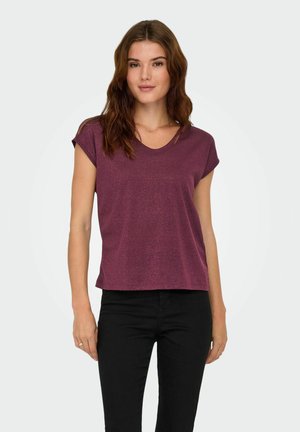 Jeune femme aux cheveux ondulés bruns portant un top violet à manches courtes et un pantalon noir, se tenant devant un fond clair uni.