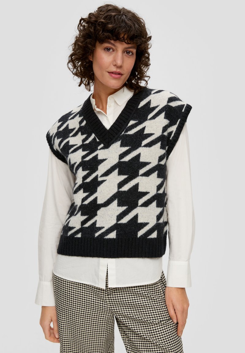 Pull sans manches noir et crème avec un motif pied-de-poule, décolleté en V et ourlet en côtes, associé à une chemise blanche et un pantalon pied-de-poule.