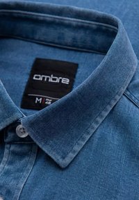 Denimhemd in Hellblau mit Knopfleiste am Kragen und einem schwarzen Etikett, auf dem "ombre" und die Größe M steht. Es zeichnet sich durch eine glatte Textur und saubere Nähte aus.