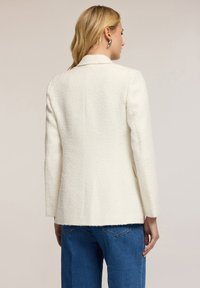 Biały żakiet o fakturowanej strukturze z dopasowanym krojem, klapami i długimi rękawami. Połączony z niebieskimi dżinsami, co ukazuje czysty, nowoczesny design.