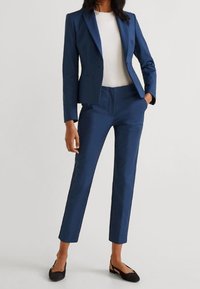 Blazer bleu marine sur mesure et pantalon assorti. Tissu lisse, coupe slim, revers crantés sur le blazer. Haut blanc en dessous. Chaussures plates noires.