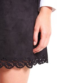 Svart mockakjol med en skålad fåll som har dekorativa utskärningar. Närbild visar en hand som greppar tyget nära den nedre kanten.