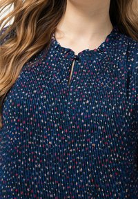 Kvinna klädd i en marinblå plisserad blus med volangkrage, knäppt nyckelhålsurringning och ett abstrakt prickmönster i röd och beige.