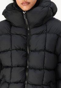 Svart puffjakke med høy krage, quiltet mønster og glidelås foran. Materialet ser glatt og blankt ut, designet for varme.
