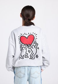 Sweat-shirt gris avec un grand cœur rouge orné de deux silhouettes en dessous, soulignées en noir, de coupe décontractée. Associé à un jean bleu clair.