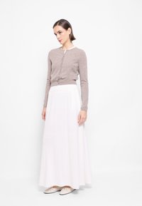 Světle hnědý pletený cardigan s kulatým výstřihem a předními knoflíky, doplněný volnou bílou maxi sukni z hladkého materiálu.