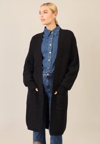 Cardigan nero in maglia con maniche lunghe, texture a costine, apertura davanti e due tasche frontali, indossato sopra una camicia di jeans e pantaloni.