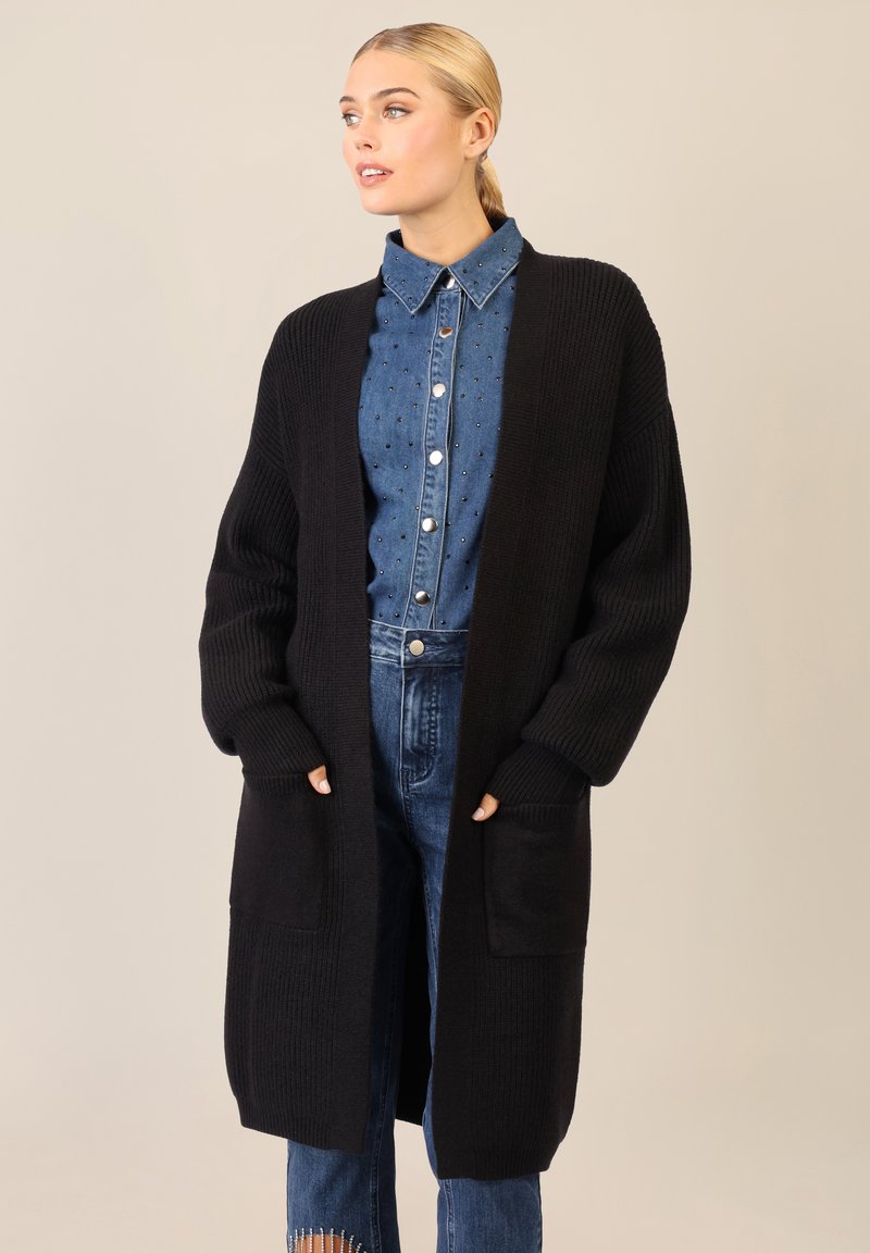 Cardigan nero in maglia con maniche lunghe, texture a costine, apertura davanti e due tasche frontali, indossato sopra una camicia di jeans e pantaloni.