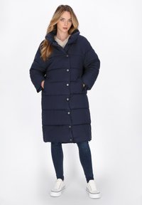 Cappotto imbottito blu navy con cappuccio, chiusura con bottoni, tasche laterali e motivo a righe orizzontali, abbinato a jeans slim e sneaker bianche.