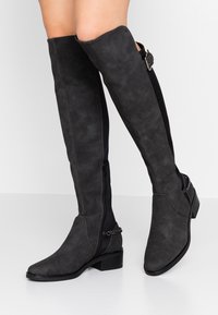 Botas negras de ante por encima de la rodilla con cremallera lateral, tacón grueso y detalle de cadena en el tobillo, con una textura mate suave.