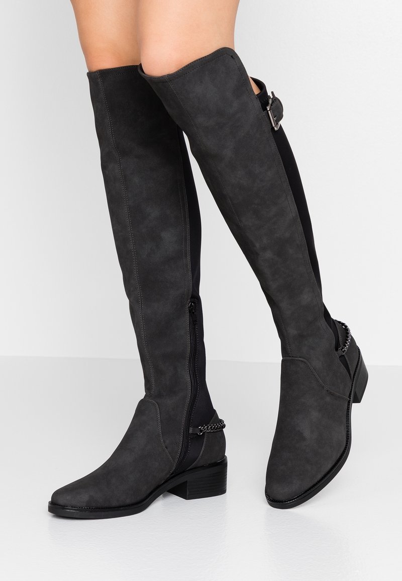 Botas negras de ante por encima de la rodilla con cremallera lateral, tacón grueso y detalle de cadena en el tobillo, con una textura mate suave.