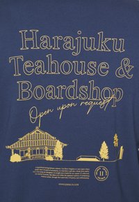 T-shirt bleu avec un texte doré : "Harajuku Teahouse & Boardshop" avec une illustration d'un bâtiment traditionnel et des arbres en dessous.