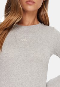 Top aderente grigio a maniche lunghe con trama a coste, scollo rotondo e sottile logo "GUESS JEANS" ricamato in colore chiaro.