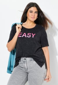 Černé tričko s krátkým rukávem a slovem "EASY" v tučném růžovém písmu. Nosené s světle šedými džíny a modrou mikinou přehozenou přes jedno rameno.