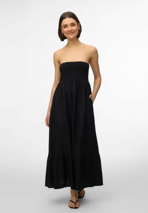 Kvinde smilende, iført sort maxikjole uden stropper med rynket liv og flæsekant, gående med hænderne i lommerne og sorte sandaler.