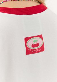 Bílá košile s červeným límečkem, kterou nosí osoba, na zádech má červenou nášivku s třešněmi a textem "Club Tropicana NAOKO".