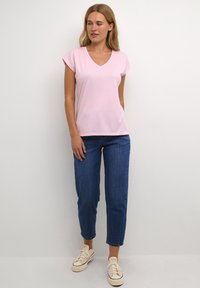 T-shirt rose à manches courtes en V associé à un jean bleu cropped et des baskets blanches, se tenant devant un fond neutre. Design simple et coupe.