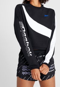 Felpa nera corta con striscia diagonale bianca, caratterizzata da un audace logo Reebok sulla manica. Abbinata a pantaloni corti neri con motivo logo.