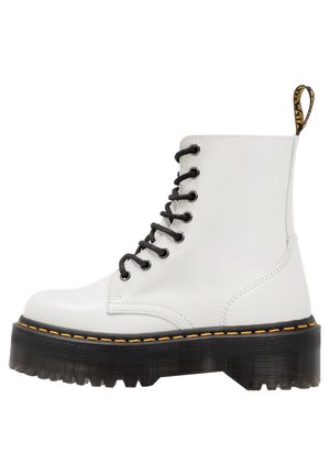 Bottes blanches à cheville en cuir lisse avec des lacets noirs, des coutures jaunes et une semelle extérieure en caoutchouc noir épaisse présentant une texture striée.