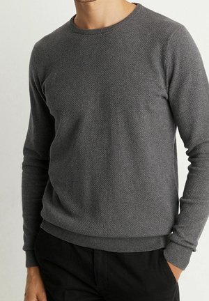 Homme portant un pull à manches longues gris foncé texturé avec des poignets et un ourlet côtelés, associé à un pantalon noir, debout avec les mains dans les poches.