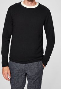 Pull noir à manches longues avec une finition texturée, doté d'un col rond blanc. Porté avec un pantalon gris à motifs, les mains dans les poches.