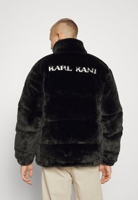Musta tekoturkki-pufferi-takki korkealla kauluksella. Takana brodeerattu "KARL KANI" -logo. Ominaisuuksiin kuuluu laatikkomainen leikkaus ja ribbaus hihansuissa. Pehmeä, teksturoitu materiaali.
