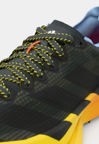 Μαύρο αθλητικό παπούτσι με κίτρινες και γκρι λεπτομέρειες. Διαθέτει υφασμένο επάνω μέρος, μαύρη λωρίδα διακόσμησης και κορδόνια σε έντονα χρώματα.