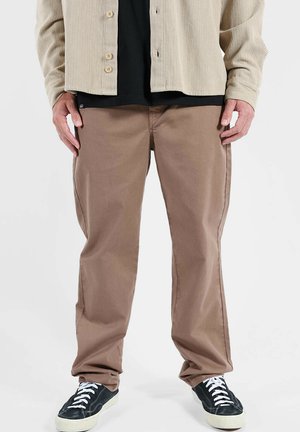 Pantalon en coton marron à coupe droite, associé à une chemise noire et une veste côtelée beige. La tenue est complétée par des baskets noires.