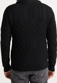 Maglione nero a maglia con motivi testurizzati e design a treccia e a rombi. Polsini e orlo a coste. Abbinato a jeans grigi.